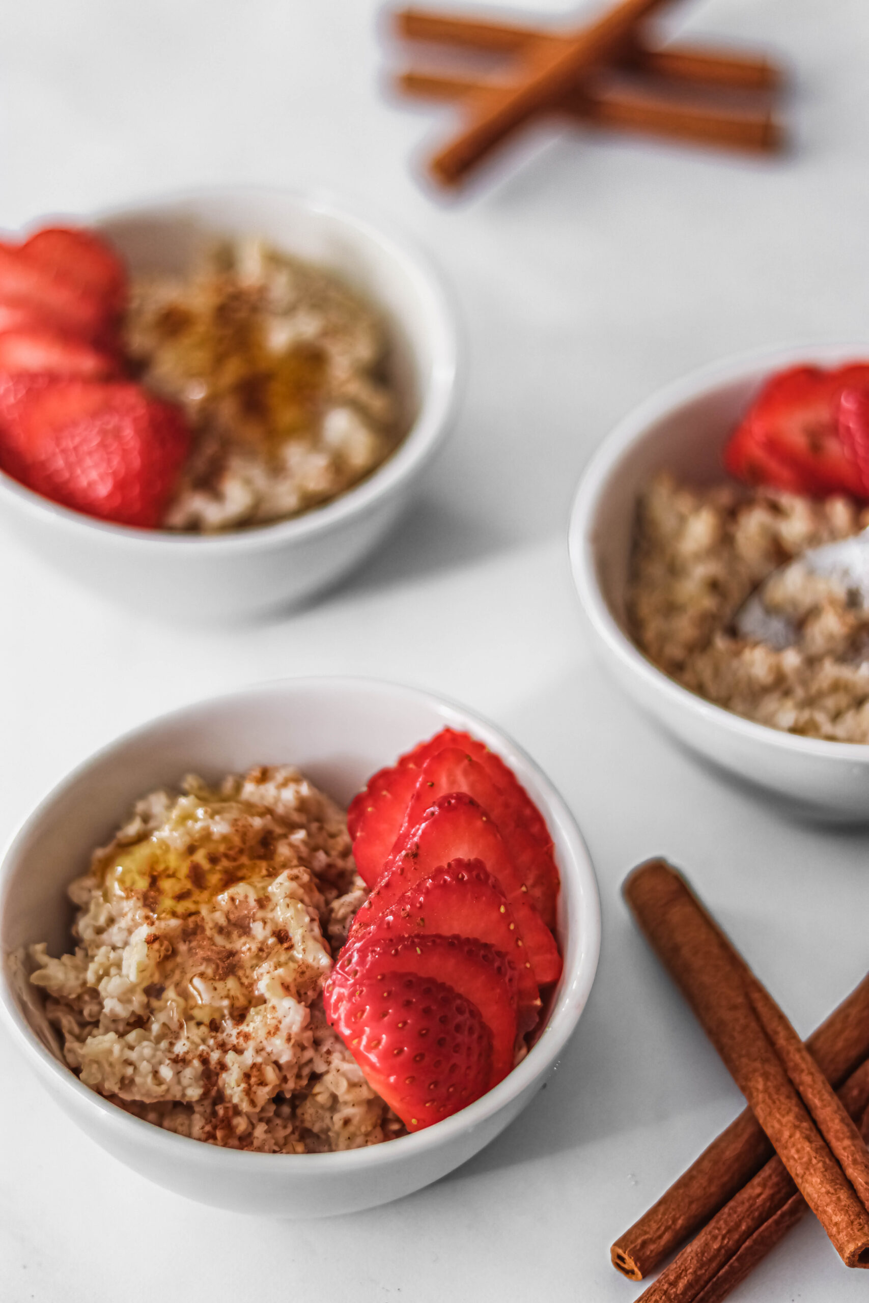 Three bowls of creamy egg white oats with freshly sliced strawberries, drizzled with honey. Full cinnamon sticks are on the side.