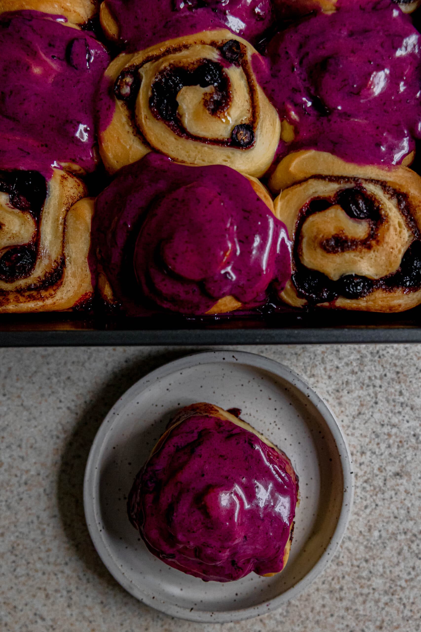 A tray of blueberry cinnamon rolls and a single roll on a plate ready to be eaten